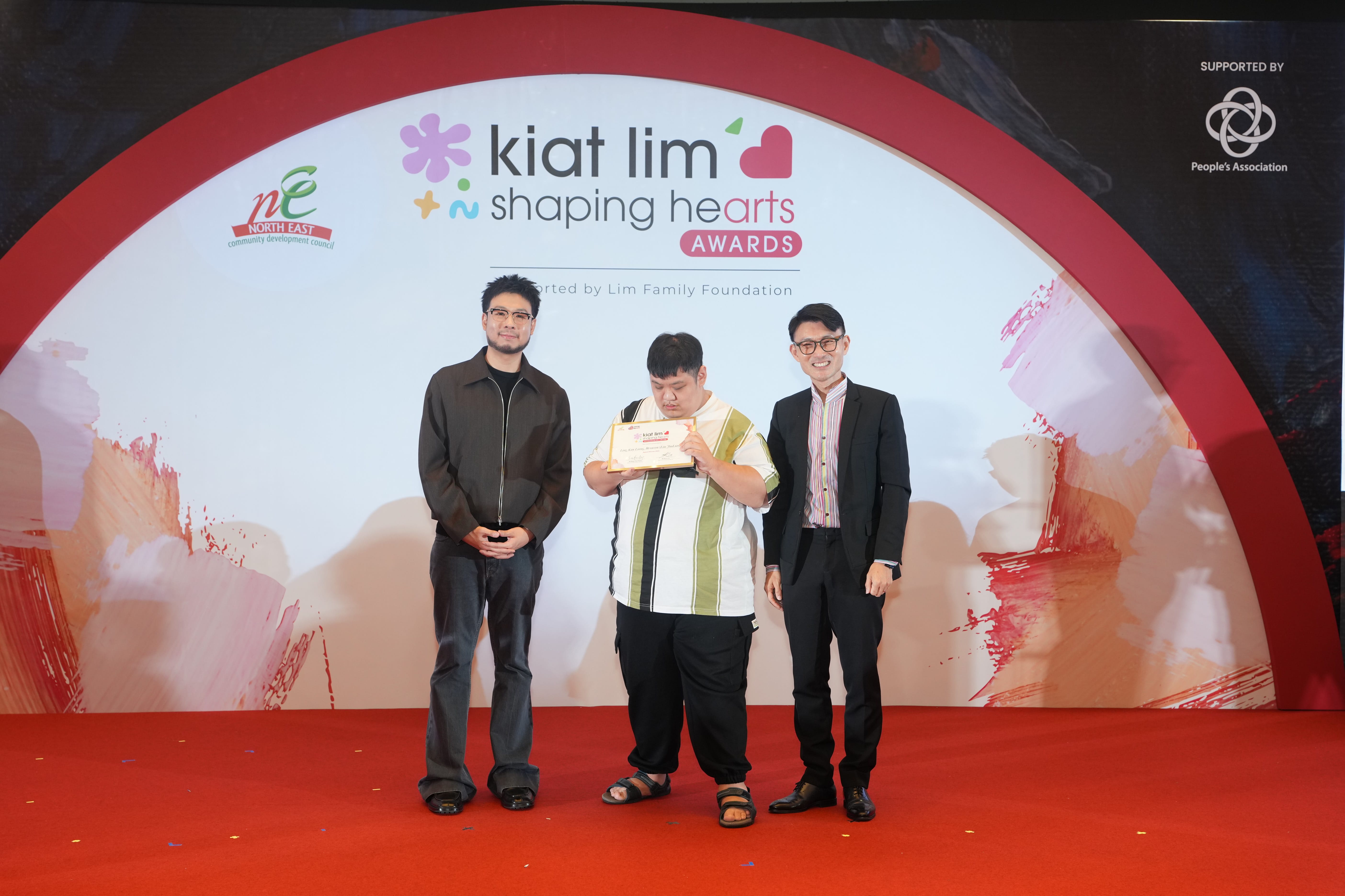 Award recipient standing on stage with the presenter, holding their certificate and posing for a group photo during the award ceremony, with a backdrop reading ‘Kiat Lim-Shaping Hearts Awards'.